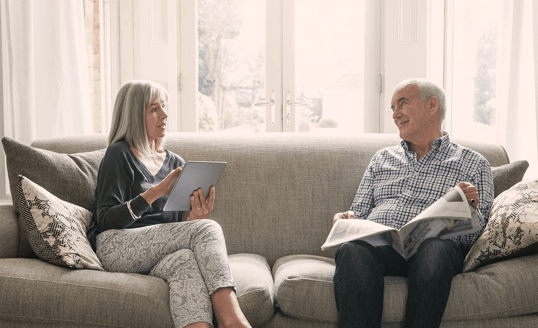 An older couple sit on a couch and talk to each other.