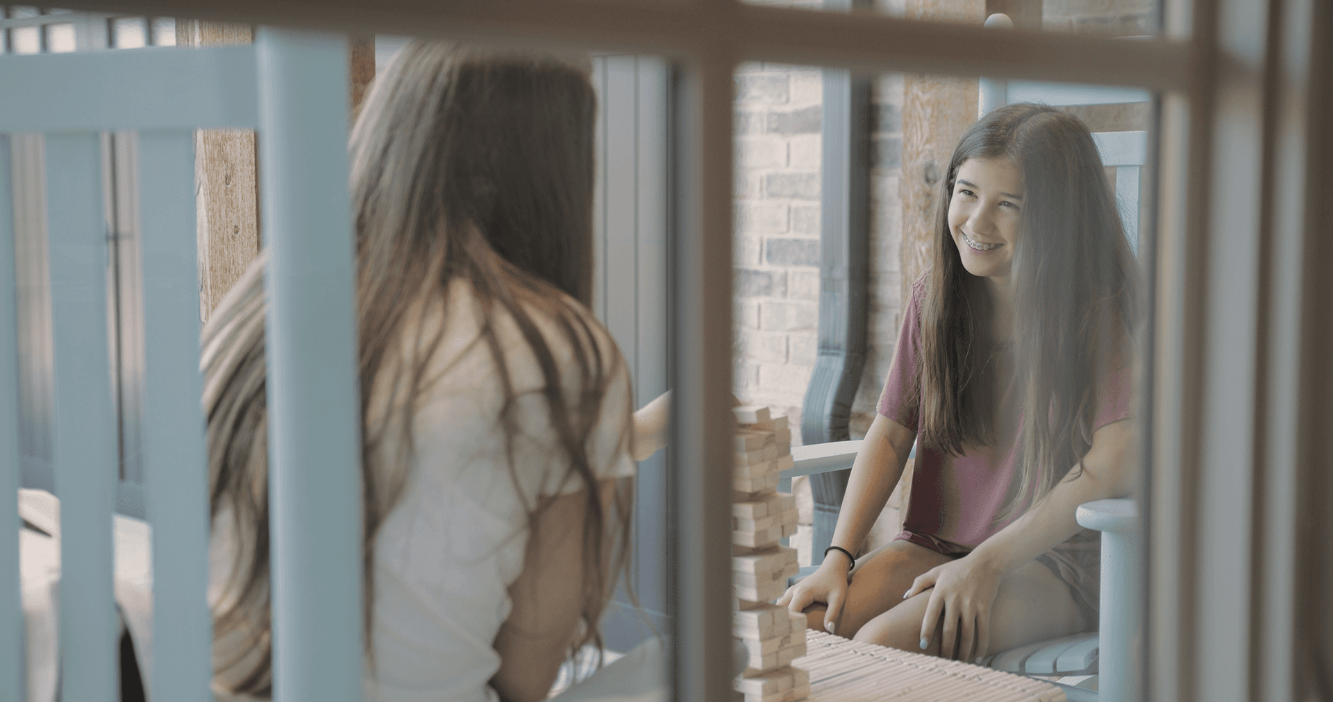 A young brunette girl with braces smiles while playing Jenga outside with another girl.