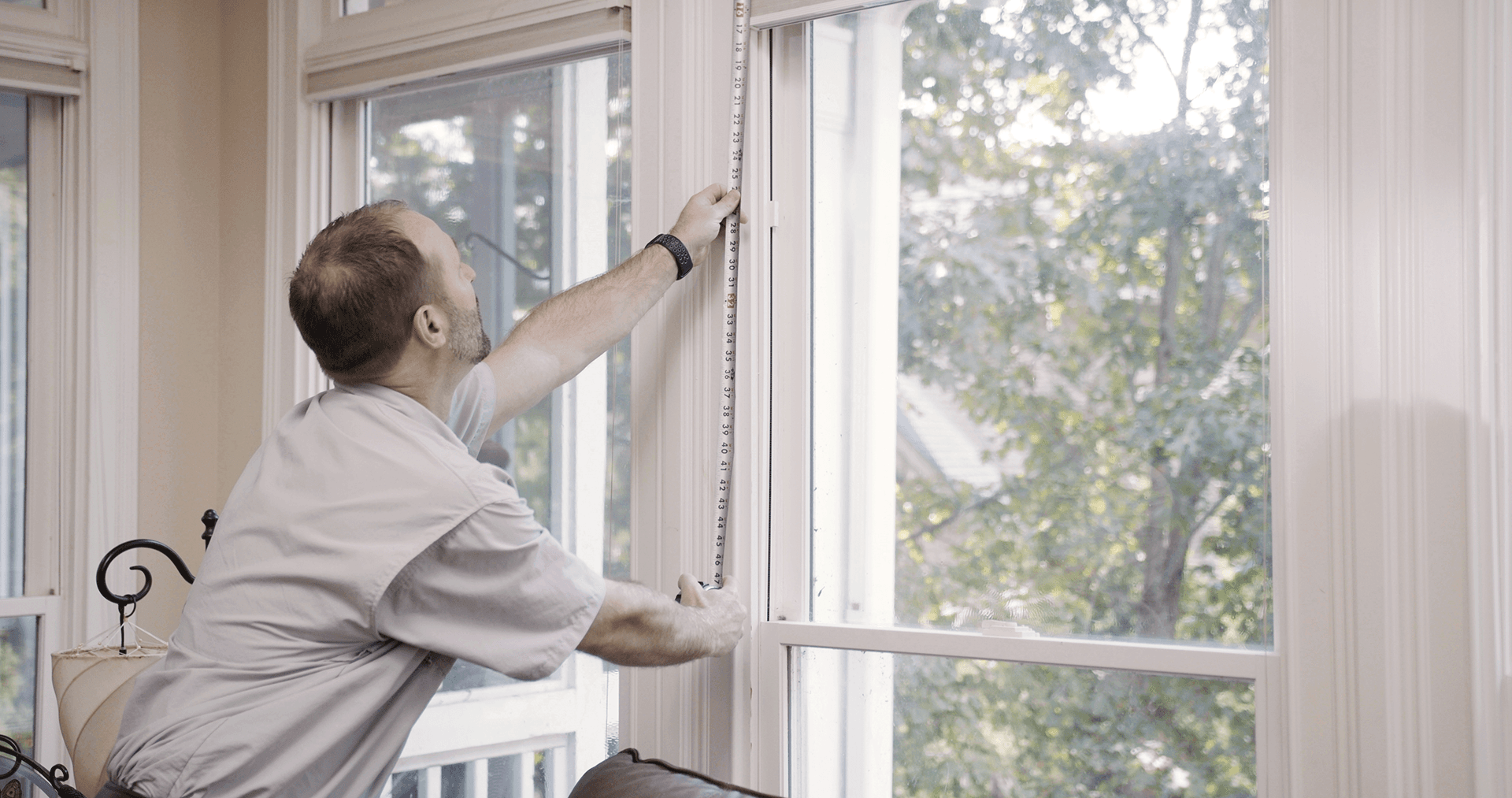 A man uses a white tape measure to measure the height of a white window inside a home.