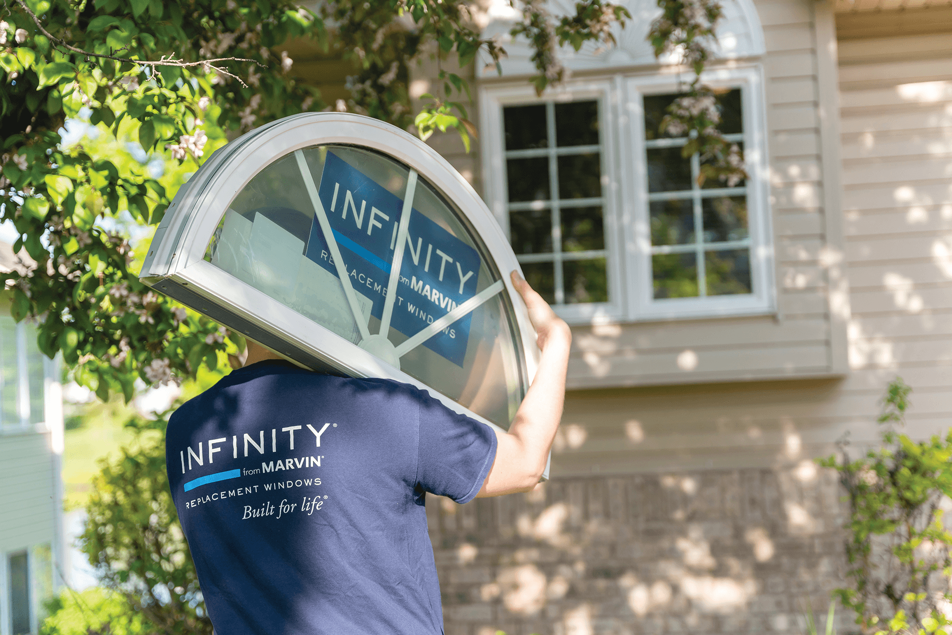 A man in a blue Infinity windows T-shirt carries an arch window on his right shoulder outside a home.